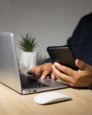 A person using a laptop computer with one hand with a phone in the other.  There is a small cactus behind the computer. 