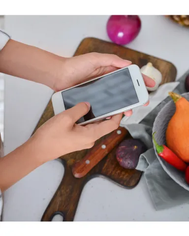 Two hands hold a cellphone over a cutting board there is a knife resting on the board and garlic and an onion nearby. 